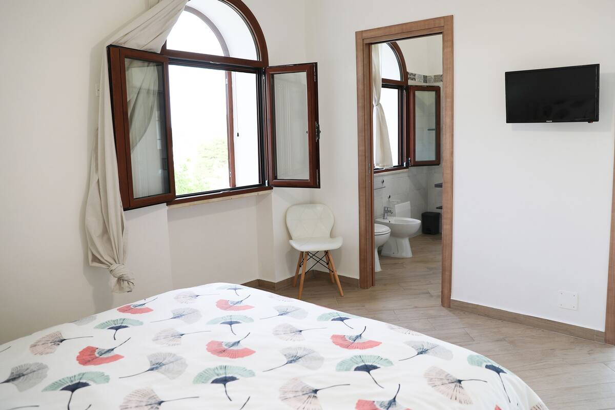 Un dormitorio con cama, ventana, espejo y televisor.