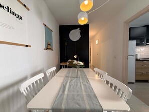 A modern dining area with a long table, white chairs, and a vase with flowers. There is a wall with a framed poster and a hanging light fixture.