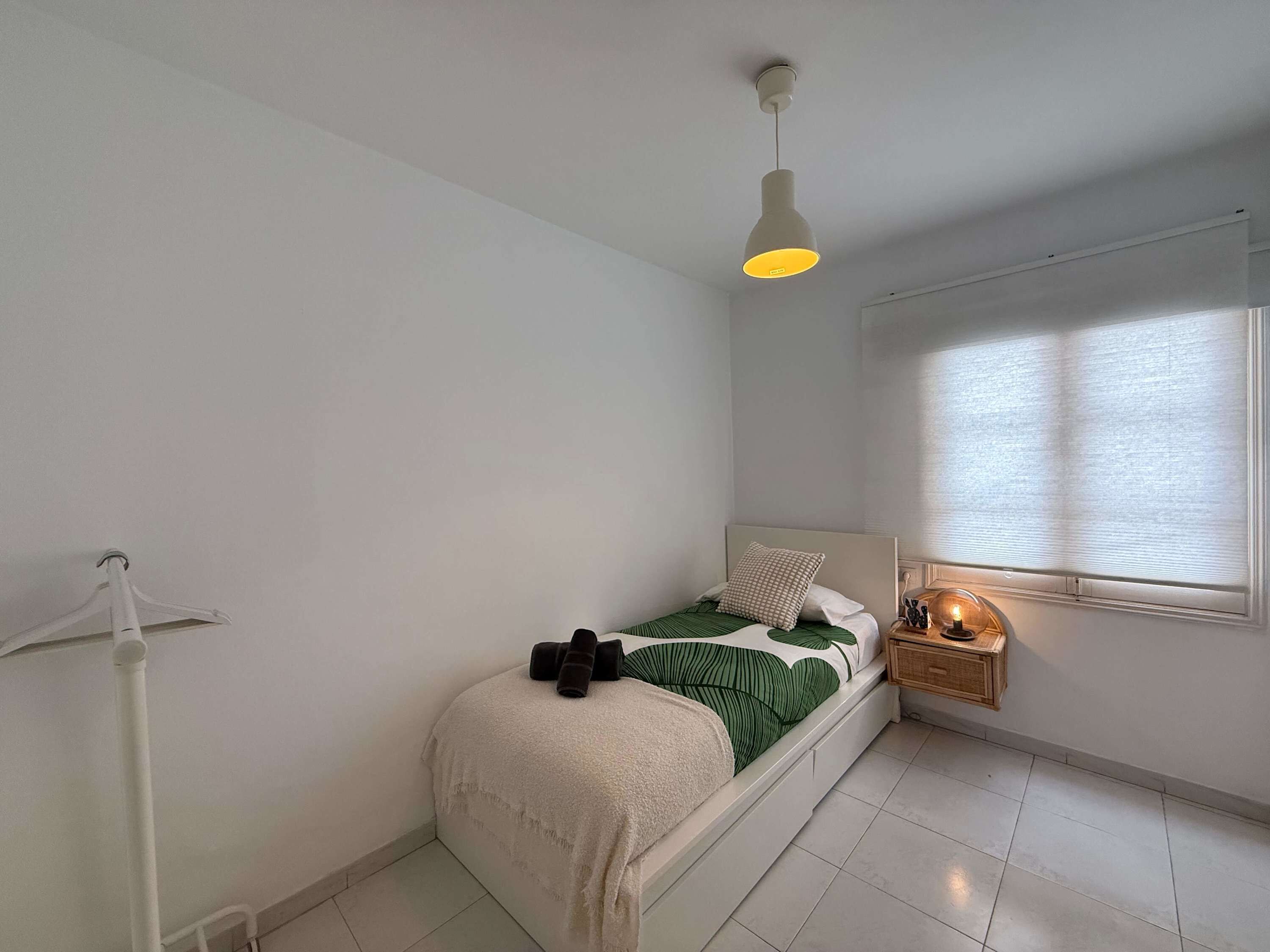 Ein Schlafzimmer mit einem Bett, einer Pendelleuchte, einem Nachttisch mit Lampe und einem Fenster mit Jalousien.