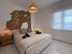 A bedroom with a large bed, a nightstand, a wall-mounted tree decoration, a hanging light fixture, and a window with blinds.