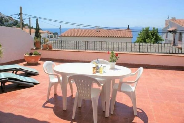 Ein runder Tisch mit vier weißen Stühlen auf einer gefliesten Terrasse, umgeben von Blumentöpfen und Blick aufs Meer.
