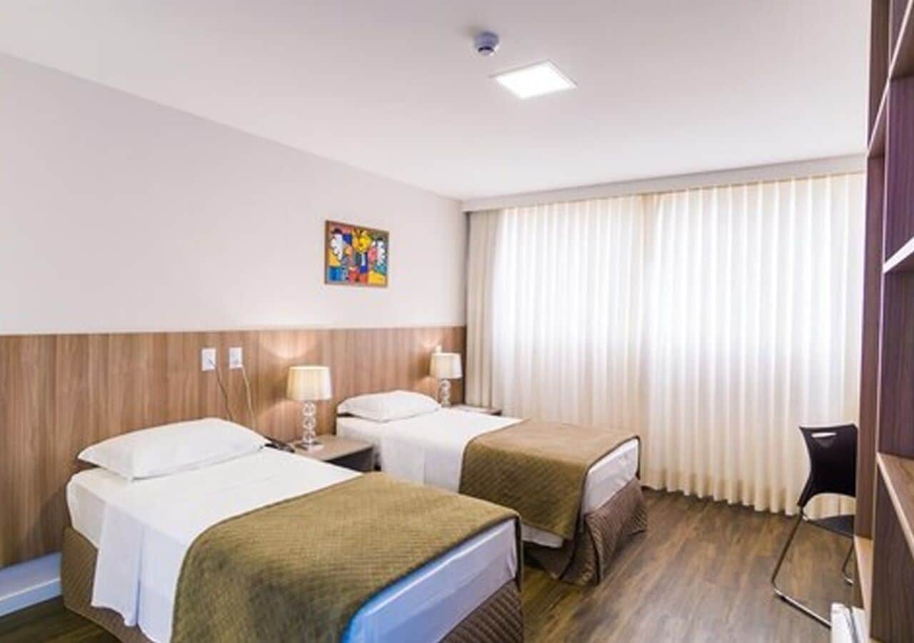 Habitació d'hotel amb dos llits, una taula, una cadira i una finestra amb cortines.