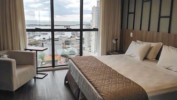 A hotel room with a large bed, a chair, a desk, and a view of the city through large windows. - Hotel 7 Itajaí by RB Hotelaria (Itajai)