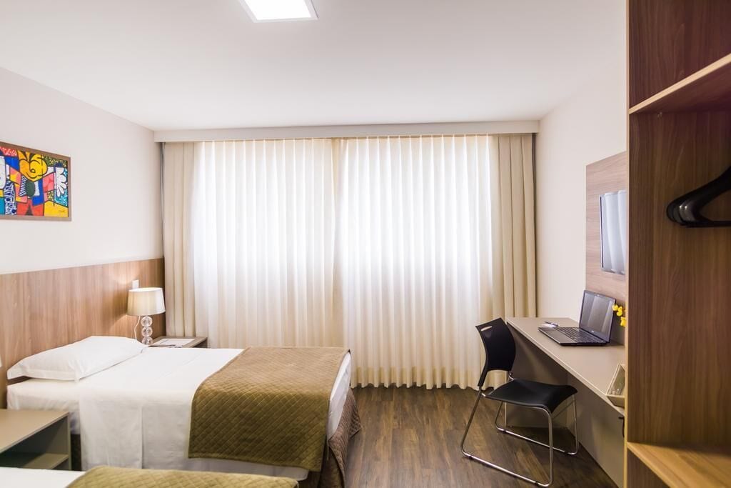 A hotel room with two beds, a desk with a laptop, a chair, and a window with curtains.