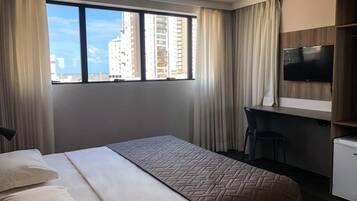A hotel room with a large bed, a desk, a TV, and a view of tall buildings.
