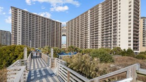 A high-rise residential building with multiple balconies, a wooden boardwalk leading to the entrance, and surrounding greenery. - 2/2 Townhome North Beach Resort- Pools, Hot tubs, (North Myrtle Beach)