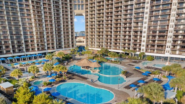 A high-rise hotel with multiple balconies, a central swimming pool area with two pools, and surrounding palm trees. - 2/2 Townhome North Beach Resort- Pools, Hot tubs, (North Myrtle Beach)