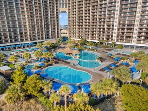 A high-rise hotel with multiple balconies, a central swimming pool area with two pools, and surrounding palm trees.