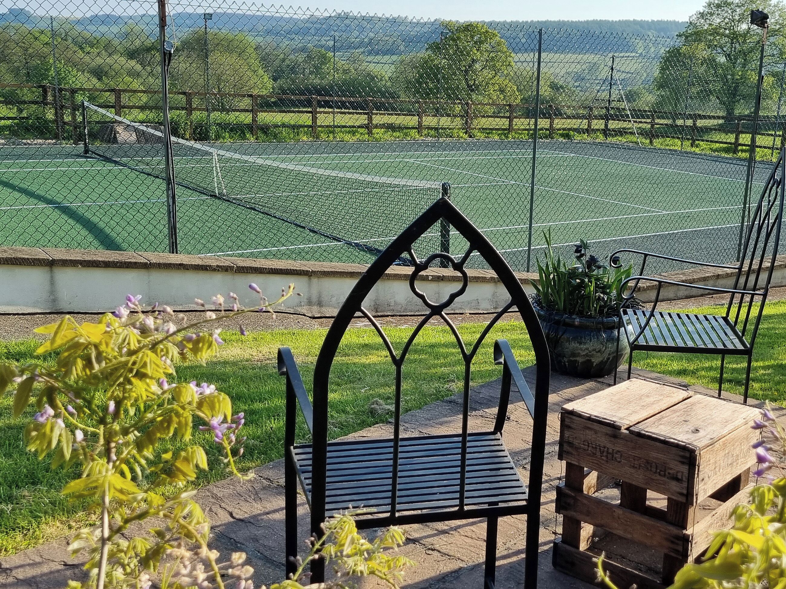 A garden with a metal bench and a wooden table, surrounded by a chain-link fence.