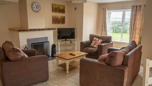 A living room with brown leather sofas, a wooden coffee table, a fireplace, a TV on a glass shelf, and a window with curtains. - House 3 Dingle Manor Remarkable 3-Bed House (Dingle)
