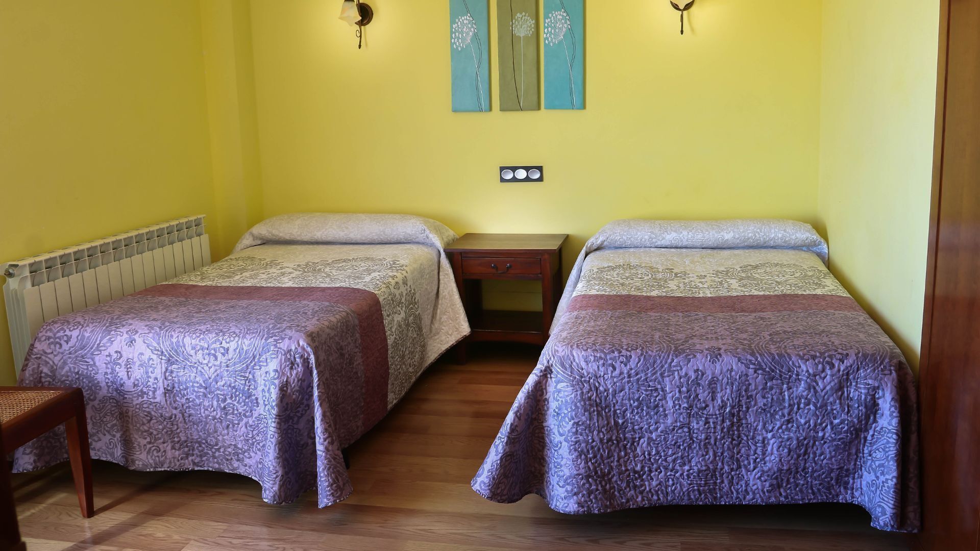 Una stanza con due letti singoli, un comodino in legno, un termosifone e una sedia.