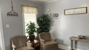 Ein Wohnzimmer mit zwei beigen Sesseln, einem Blumentopf, einem kleinen Holztisch und einem Schild mit der Aufschrift 'farmhouse'.