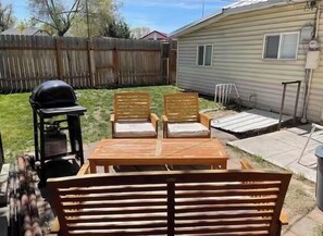 A backyard with a wooden table and two chairs, a grill, and a covered patio area.