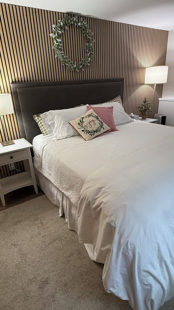 Una cama bien hecha con un cabecero oscuro, ropa de cama blanca y almohadas decorativas. Se ve una mesita de noche con una lámpara a la izquierda.