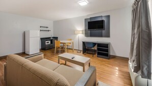 A modern living room with a sofa, a coffee table, a TV mounted on the wall, a dining area with chairs, and a kitchen area with a refrigerator and stove. - Studio 6 Laredo, TX - North I-35 (Laredo)