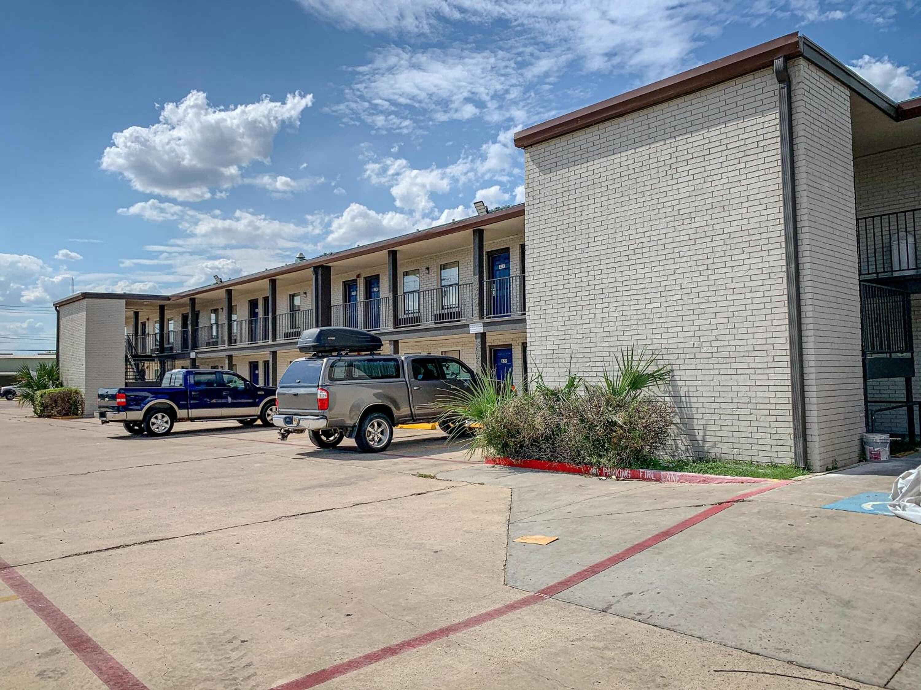 Un motel à la façade en briques, doté de nombreux balcons et d’un parking où sont stationnés plusieurs véhicules.