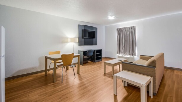 A modern living room with a wooden floor, a sofa, a small table, a desk with a chair, and a TV mounted on the wall. - Studio 6 Laredo, TX - North I-35 (Laredo)
