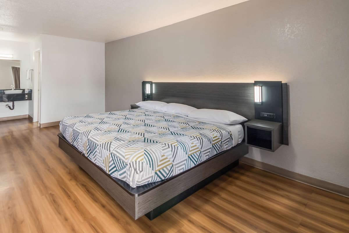 A modern bedroom with a wooden floor, a bed with patterned bedding, a nightstand, and a wall-mounted light fixture.
