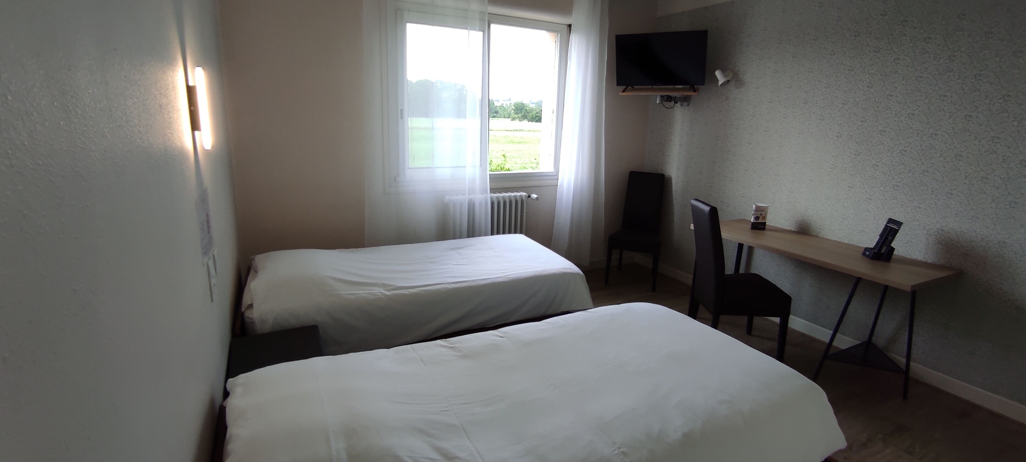 Habitación de hotel con dos camas individuales, un escritorio con silla, un televisor montado en la pared y una ventana con cortinas.