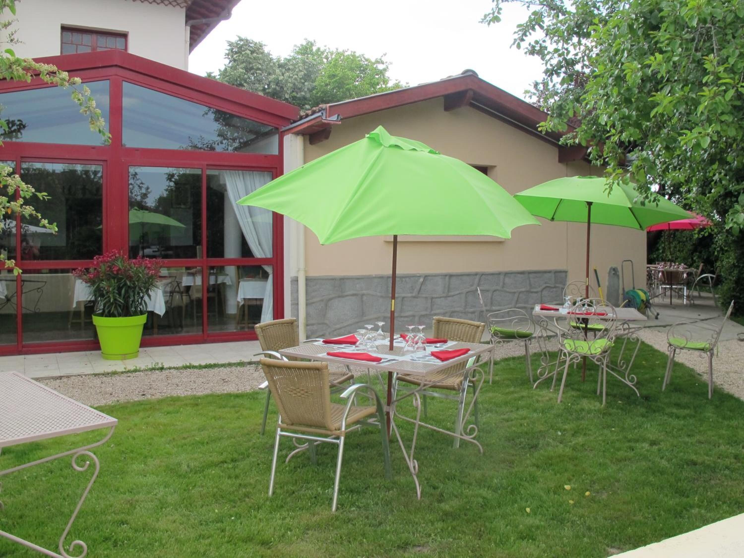 An outdoor dining area with tables and chairs, green umbrellas, and a building with large windows and a red frame.
