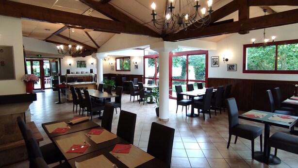 Restaurante con techos altos, vigas a la vista y grandes ventanales que ofrecen vistas al verdor. Las mesas están dispuestas con servilletas rojas y cubiertos.