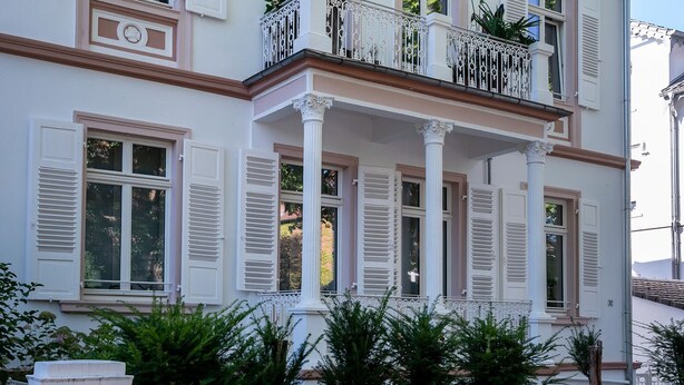Een meerhoog gebouw met witte muren en roze accenten, met meerdere ramen voorzien van luiken, een balkon met decoratieve relingen en een verzorgde tuin.