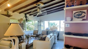 A spacious living room with a white sofa, a coffee table, and a lamp. There are chairs, a plant, and a shelf with decorative items.
