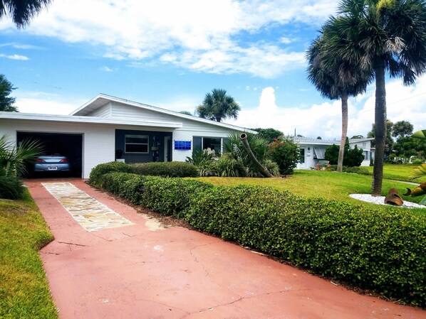 Una casa moderna con garaje, entrada pavimentada y jardines bien cuidados, incluyendo setos y palmeras.