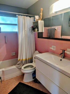 Un baño con bañera, inodoro y lavamanos. Cuenta con una cortina de ducha, un espejo y un estante con artículos de tocador.