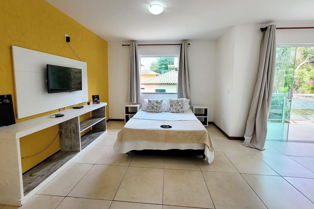 Habitación de hotel con cama, televisor montado en la pared, mesita de noche pequeña y ventana con cortinas.