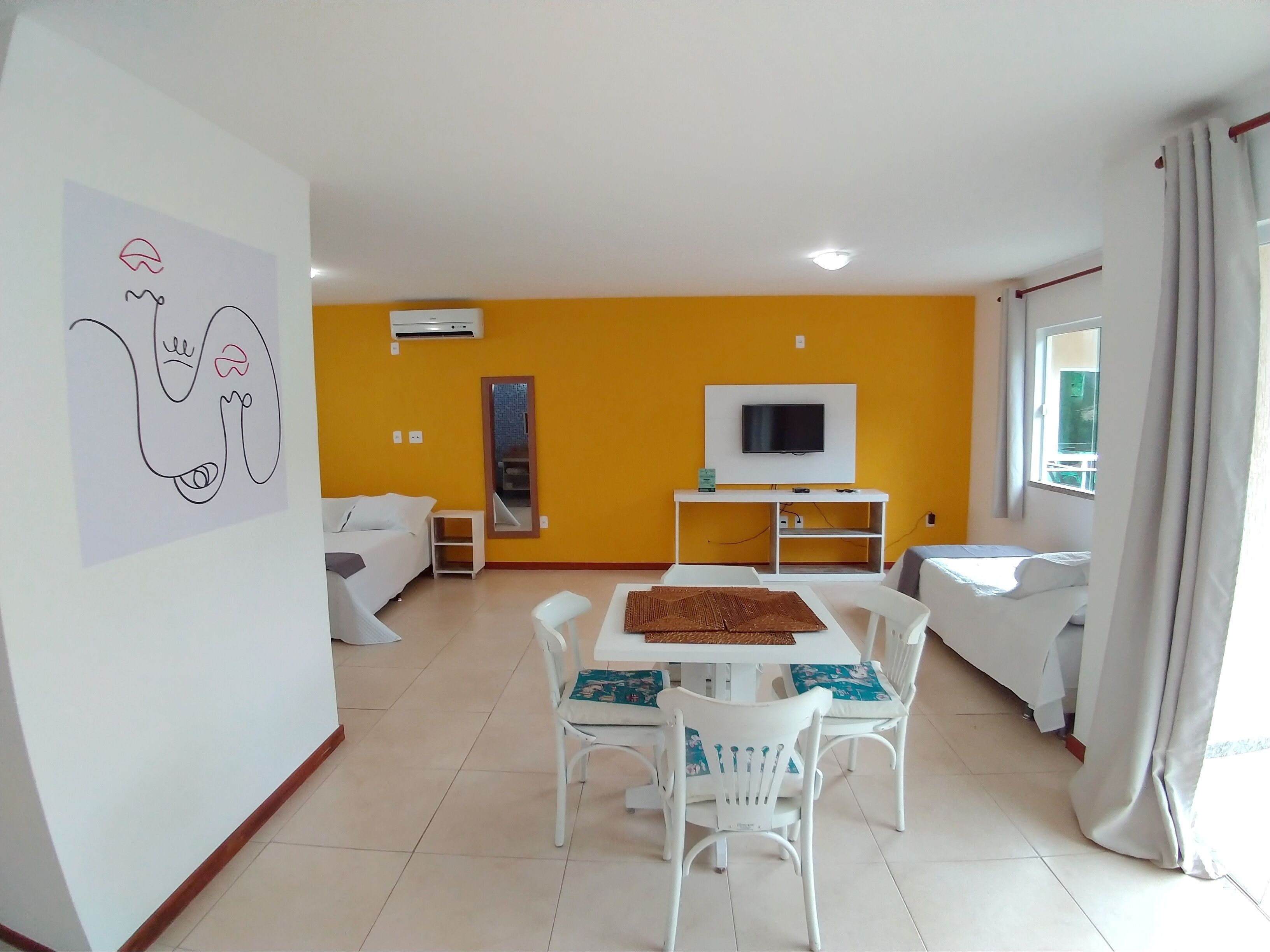 A modern living room with a yellow accent wall, a flat-screen TV, a dining table with chairs, and a white sofa.