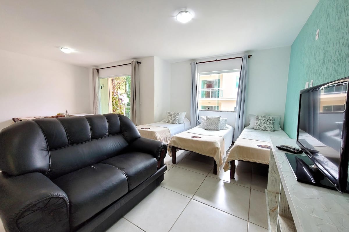 Una sala de estar con un sofá de cuero negro, dos camas individuales con ropa de cama blanca, un televisor sobre un mueble bajo y una ventana con cortinas.