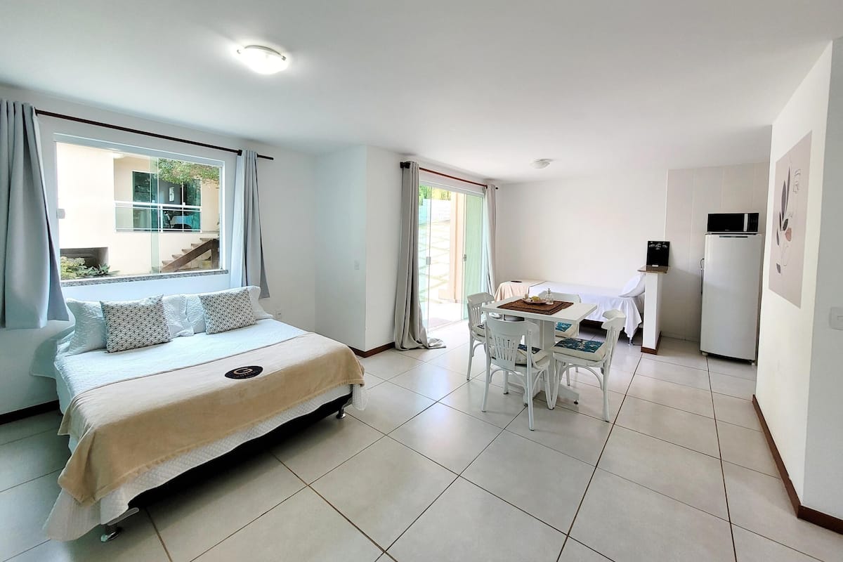 Un dormitorio con cama, una mesa de comedor con sillas, un refrigerador y una ventana con cortinas.