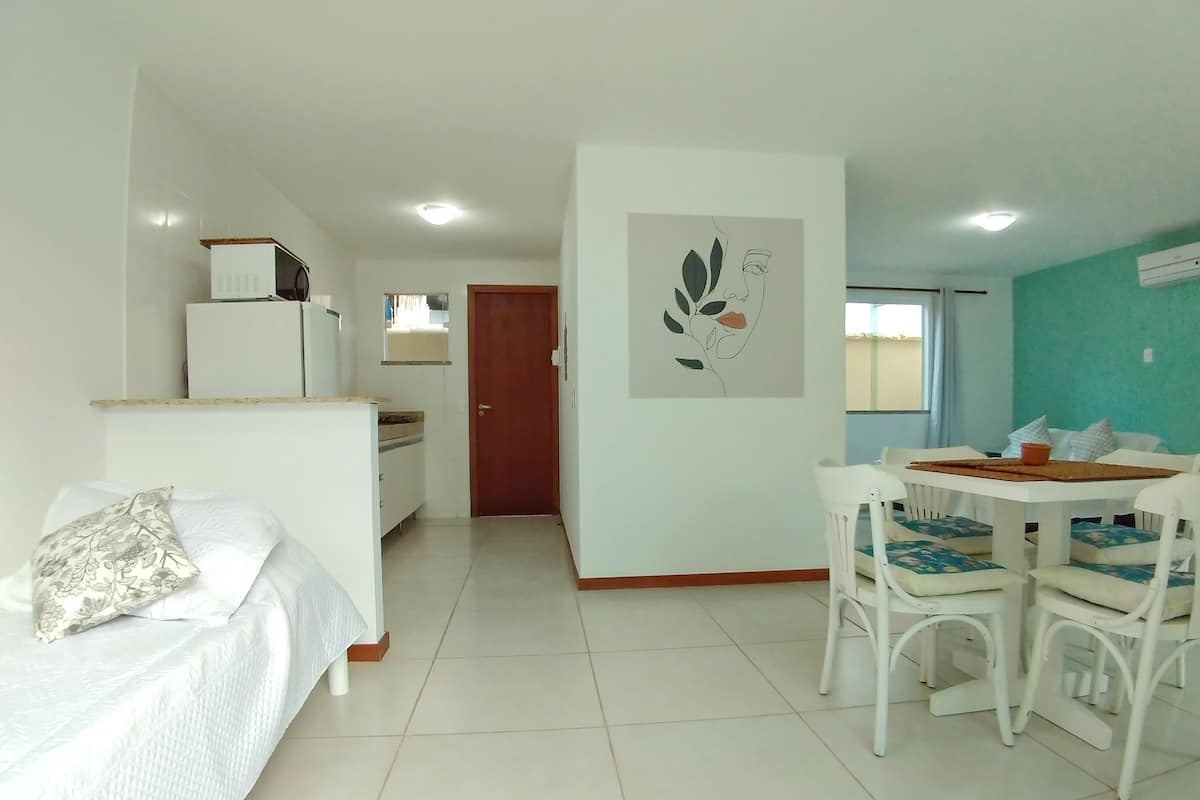 Una habitación de hotel con una cama, una kitchenette pequeña, una zona de comedor con una mesa y sillas, y una puerta que da a otra habitación.