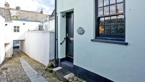 Une ruelle étroite pavée de pierres menant à un bâtiment blanc doté d’une porte et d’une fenêtre noires.