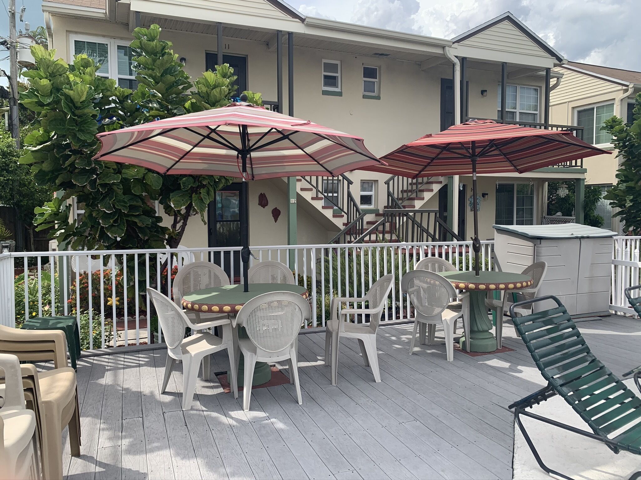 Une terrasse extérieure couverte, avec des chaises et des tables en plastique blanc, deux parasols rouges et une clôture blanche.