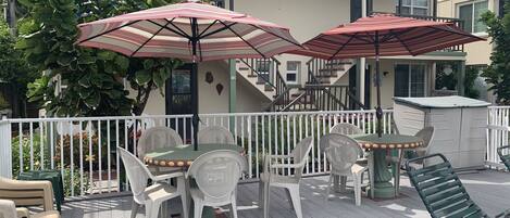 A covered outdoor patio with white plastic chairs and tables, two red umbrellas, and a white fence.