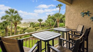 A balcony with two tables and chairs, overlooking a beach with palm trees and a clear sky.