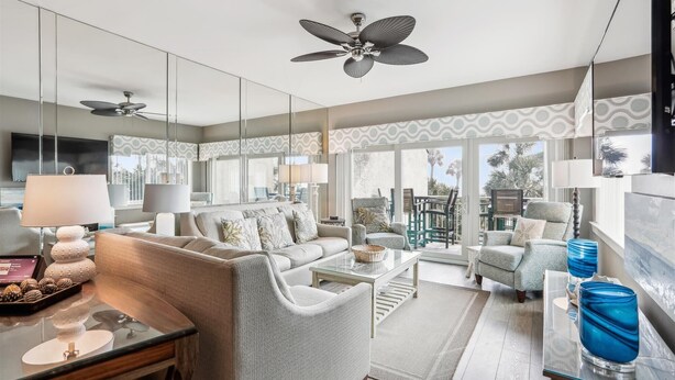 A modern living room with a sofa, armchairs, a coffee table, and a TV. There are ceiling fans, a large window with a view, and a patterned wall.