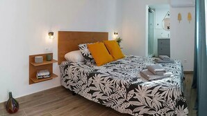 Quarto com cabeceira de madeira, colcha de cama estampada em preto e branco, duas almofadas amarelas, mesa de cabeceira com um vaso e um espelho na parede.
