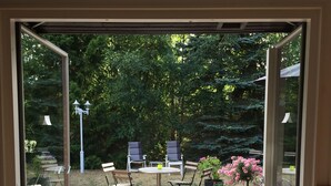 Una terraza de madera con una mesa y sillas, un tapete rojo y vistas a un jardín con árboles y flores.