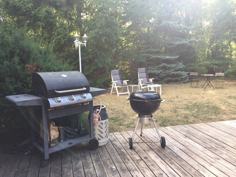 Una parrilla para barbacoa y una parrilla portátil en una terraza de madera, con sillas y mesas al fondo.