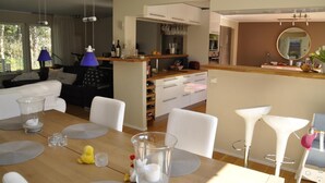 Una sala de estar moderna con una mesa de comedor de madera, sofás blancos y una cocina con armarios blancos y una barra.