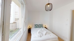 A bedroom with a bed, two bedside lamps, a hanging light fixture, and a view through a window.