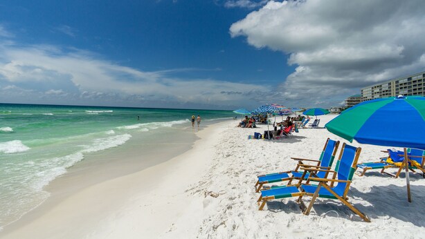 A beach with white sand, clear turquoise water, and a few people walking. There are blue and green umbrellas and wooden chairs lined up on the sand.