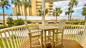 A wooden deck with a table and two chairs, surrounded by white railings and palm trees.