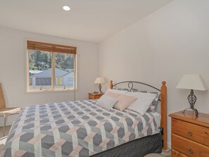 Un dormitorio con cama, dos mesitas de noche, una silla y ventana con vista a casas.