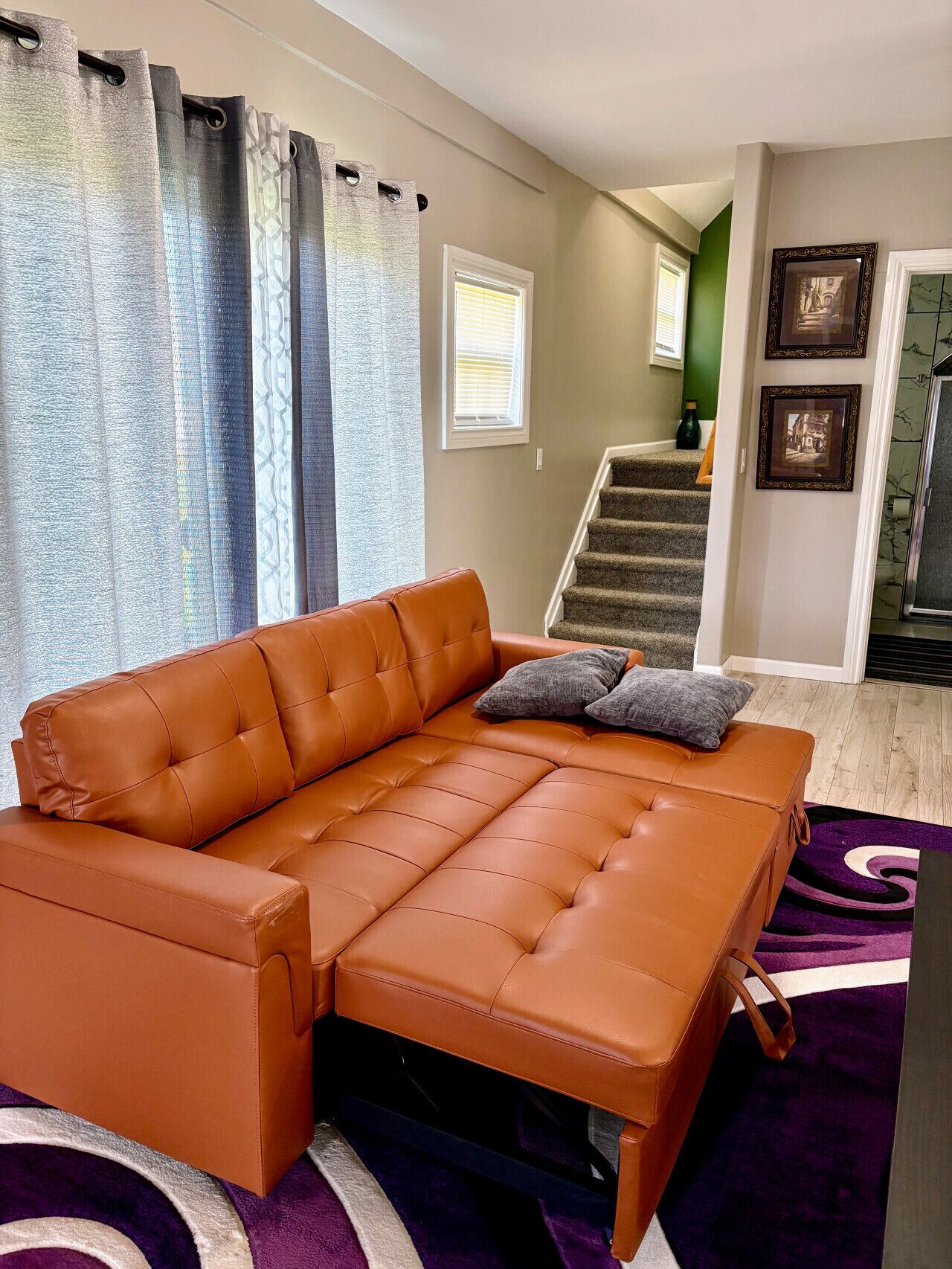 A living room with a brown leather sofa, a purple rug, and a staircase leading to a second floor.