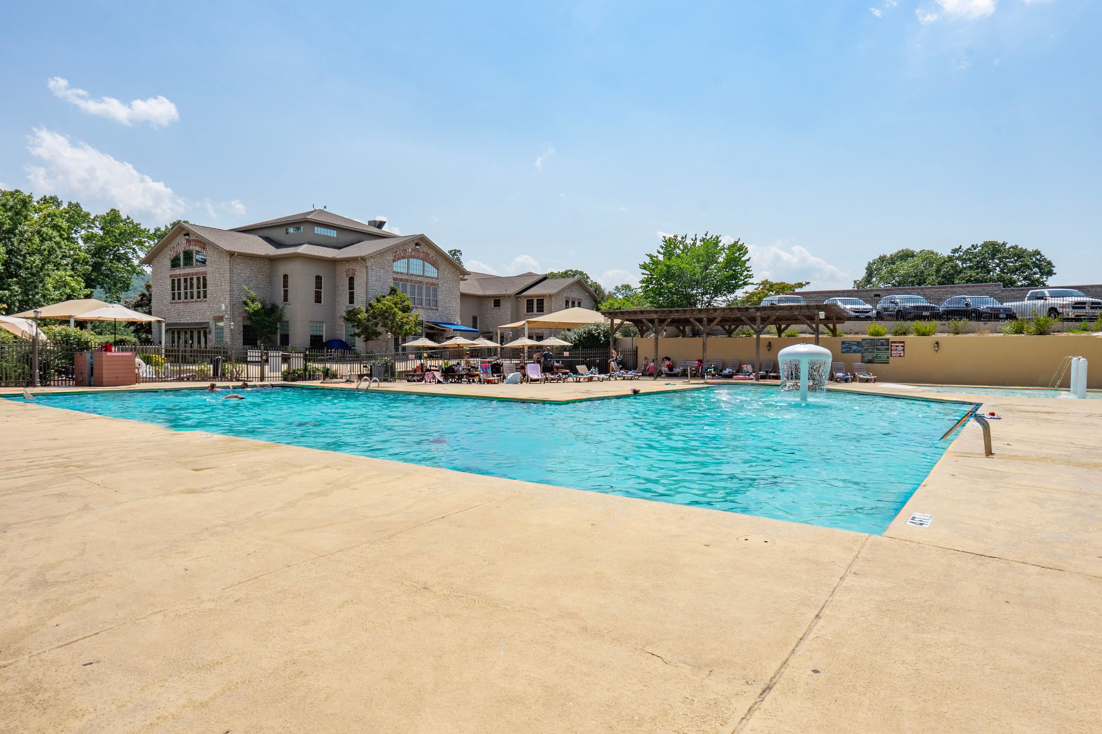 A swimming pool with lounge chairs and umbrellas, a stone building with a sign, and a parking area with cars.