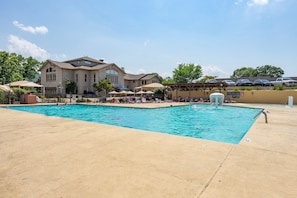 A swimming pool with lounge chairs and umbrellas, a stone building with a sign, and a parking area with cars.